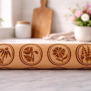 Bright, airy kitchen scene featuring a wooden embossed rolling pin with floral patterns placed on a marble countertop. The background is softly blurred, showing a white ceramic pot filled with fresh flowers, a wooden cutting board, and minimal kitchen elements. Natural light enhances the warm tones of the wood and highlights the intricate engraved details, creating a calm, elegant, and inviting atmosphere.