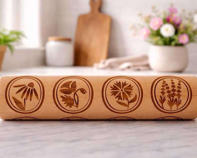 Bright, airy kitchen scene featuring a wooden embossed rolling pin with floral patterns placed on a marble countertop. The background is softly blurred, showing a white ceramic pot filled with fresh flowers, a wooden cutting board, and minimal kitchen elements. Natural light enhances the warm tones of the wood and highlights the intricate engraved details, creating a calm, elegant, and inviting atmosphere.