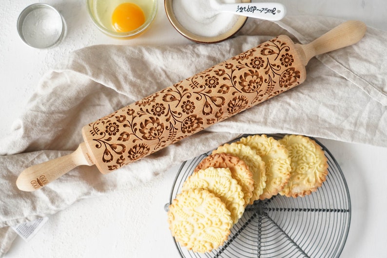 Wooden rolling pin with decorative design of polish folk traditional flowers. A well baked cookies presenting the effects of embossing are lying on a metal trivet  in front of it.