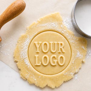 May include: A round cookie stamp pressed into a piece of light beige dough, creating the embossed text "YOUR LOGO". The dough is dusted with white flour and positioned next to a wooden rolling pin and a circular metal cookie cutter.