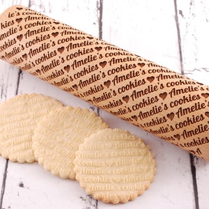 May include: A wooden rolling pin with the text "Amelie's cookies" engraved on it. Three cookies with the same text imprinted on them are in the foreground.