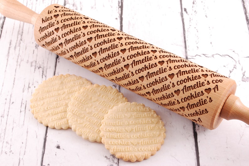 May include: A wooden rolling pin with the text "Amelie's cookies" engraved on it. Three cookies with the same text imprinted on them are in the foreground.
