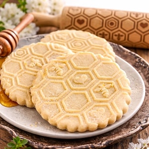 Può includere: Primo piano di tre biscotti rotondi e chiari con un motivo a nido d'ape e disegni di api e cuori. I biscotti sono su un piatto decorativo, con un mattarello di legno e un misurino di miele sullo sfondo. I biscotti hanno un bordo smerlato.