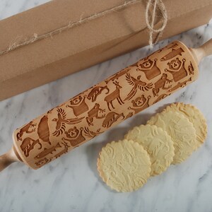 A beautifully styled product scene featuring a wooden embossed rolling pin with intricate animal patterns, placed diagonally on a soft, light background. Next to it lies a kraft gift box tied with natural twine, enhancing the rustic yet elegant aesthetic. In the foreground, several round cookies display detailed imprints from the rolling pin, showcasing real baking results. Delicate dried flowers add a refined, minimal touch, while soft lighting and gentle shadows create inviting composition.