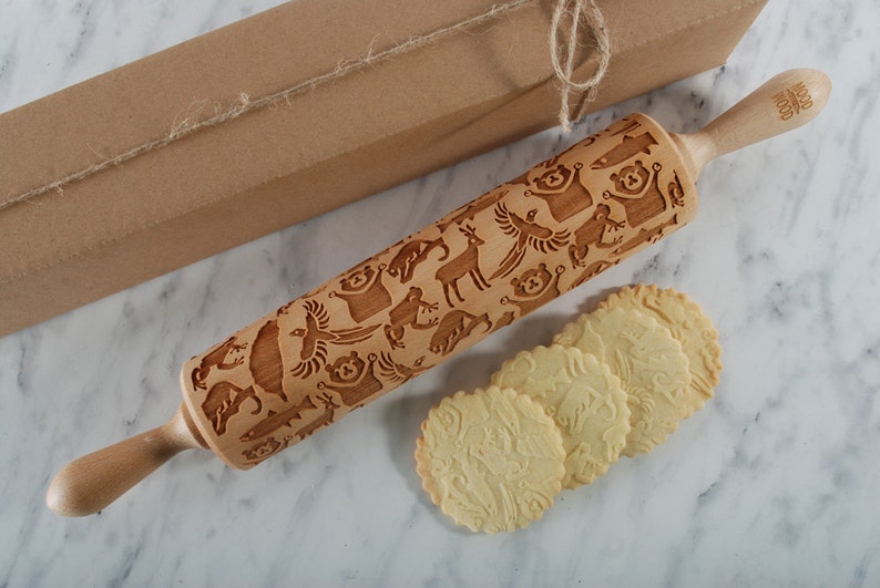 A beautifully styled product scene featuring a wooden embossed rolling pin with intricate animal patterns, placed diagonally on a soft, light background. Next to it lies a kraft gift box tied with natural twine, enhancing the rustic yet elegant aesthetic. In the foreground, several round cookies display detailed imprints from the rolling pin, showcasing real baking results. Delicate dried flowers add a refined, minimal touch, while soft lighting and gentle shadows create inviting composition.
