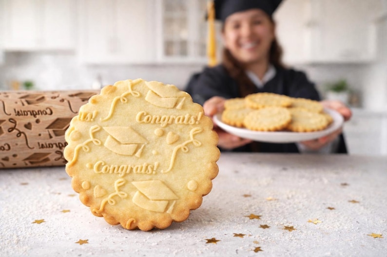 High-quality product photo showing a wooden engraved rolling pin with a detailed graduation-themed pattern, including caps, hearts, and “Congrats” text. A realistic magnifying glass highlights the exact same engraving, revealing deep, precise carving and natural wood grain texture. Surrounding baking elements like eggs, flour, and freshly baked embossed cookies create a warm, artisanal kitchen scene. Soft natural lighting and shallow depth of field emphasize craftsmanship and premium quality.