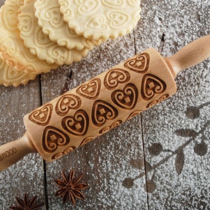 May include: A wooden rolling pin with a heart-shaped design engraved on the surface. The rolling pin is on a wooden surface with white powder, and there are cookies in the background.