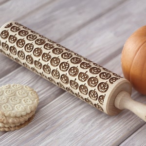 Wooden rolling pin with halloween pumpkins design. A orange pumpkin is standing behind it. A pile of well baked cookies with embossed design is in front of a rolling pin