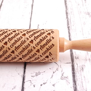 May include: A wooden rolling pin with a handle and the text "Amelie's cookies" engraved on the surface. The rolling pin is on a white wooden surface.