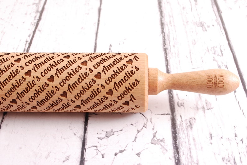May include: A wooden rolling pin with a handle and the text "Amelie's cookies" engraved on the surface. The rolling pin is on a white wooden surface.