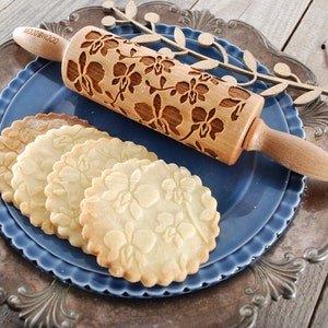 May include: A wooden rolling pin with an orchid design is shown on a blue plate with four cookies that have been rolled with the pin. The cookies are on a silver plate.