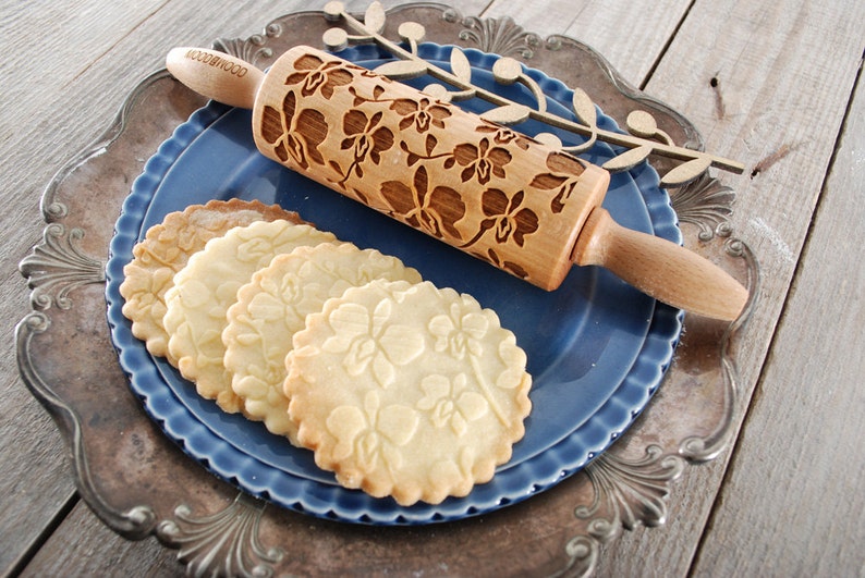 May include: A wooden rolling pin with an orchid design is shown on a blue plate with four cookies that have been rolled with the pin. The cookies are on a silver plate.