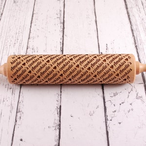 May include: A wooden rolling pin with a brown handle and a natural wood body. The body is engraved with the text "Amelie's cookies" repeated many times. The rolling pin is on a white wooden surface.