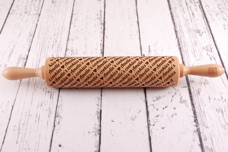 May include: A wooden rolling pin with a brown handle and a natural wood body. The body is engraved with the text "Amelie's cookies" repeated many times. The rolling pin is on a white wooden surface.