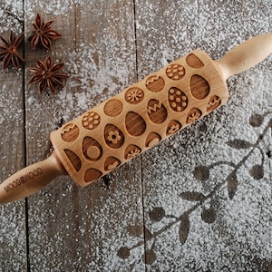 Peut inclure: Un rouleau à pâtisserie en bois avec un motif gravé d'œufs de Pâques. Le rouleau à pâtisserie est sur une surface en bois recouverte de poudre blanche.