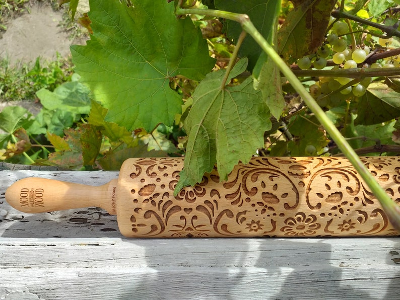 Wooden embossing rolling pin with moodforwood logo on the handle, embossing design is rosemaling, norwegian folk. The pin is lyingbehind the wine leaes on grey rustic board