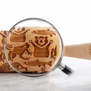Close-up product image of a wooden embossed rolling pin featuring detailed animal engravings, placed on a smooth, light surface. A realistic magnifying glass overlays the center, enlarging the exact same pattern to highlight the depth and precision of the carving. The zoomed area clearly reveals natural wood grain and approximately 3 mm deep engravings. A partially visible cookie appears on the left, reinforcing real baking results, while soft lighting and shadows create  premium aesthetic.