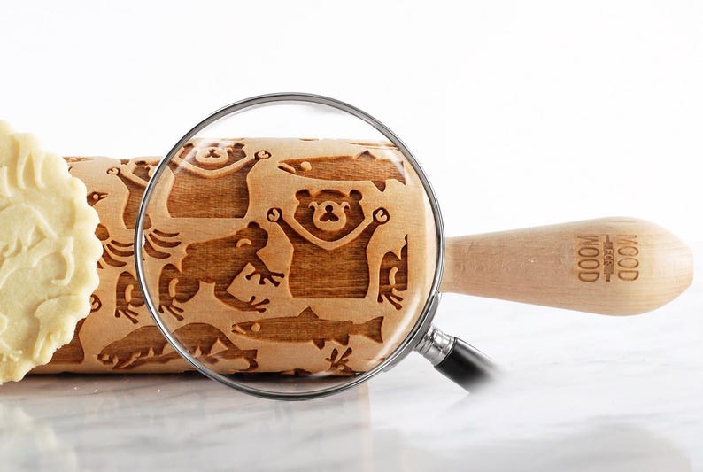 Close-up product image of a wooden embossed rolling pin featuring detailed animal engravings, placed on a smooth, light surface. A realistic magnifying glass overlays the center, enlarging the exact same pattern to highlight the depth and precision of the carving. The zoomed area clearly reveals natural wood grain and approximately 3 mm deep engravings. A partially visible cookie appears on the left, reinforcing real baking results, while soft lighting and shadows create  premium aesthetic.