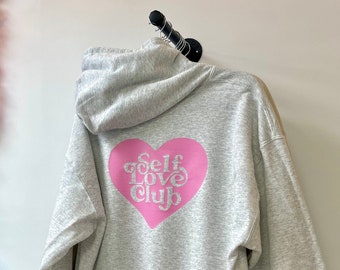 May include: Gray hooded sweatshirt with a pink heart graphic that says "Self Love Club" on the back.