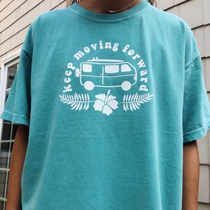 May include: A teal blue t-shirt with a white graphic design of a vintage van with surfboards on top. The graphic also includes the text "Keep moving forward" and a hibiscus flower.
