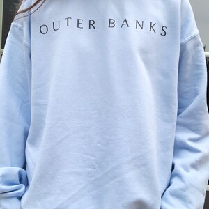 May include: A light blue sweatshirt with the text "OUTER BANKS" printed in black on the front.