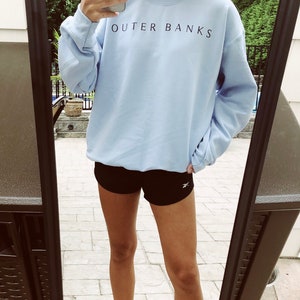 May include: A light blue sweatshirt with the text "OUTER BANKS" printed in black on the front. The sweatshirt is being worn by a person wearing black athletic shorts.