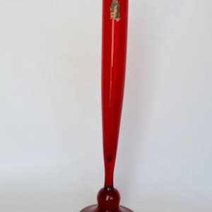 May include: A tall, slender red glass vase with a flared base and a bulbous midsection. The vase has a long, cylindrical neck and a small sticker near the top. The vase is approximately 30 cm tall.