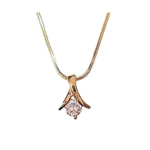 May include: A gold-toned necklace with a pendant featuring a clear, round gemstone. The pendant has a stylized, curved design that cradles the gem. The chain is a delicate, snake-style chain. The necklace is set against a white background.