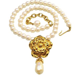 May include: A white pearl necklace with a gold flower pendant and a single pearl drop. The necklace has a clasp closure and a chain extender.