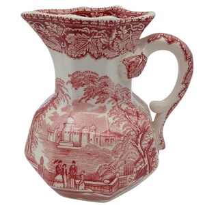 May include: A white and red ceramic pitcher with a detailed landscape design. The pitcher features a handle and a unique, scalloped rim. The red design includes trees, buildings, and figures, creating a classic, vintage aesthetic.
