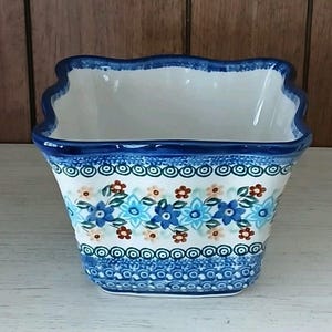 May include: A square ceramic planter with a scalloped rim, featuring a blue floral pattern on a white background. The planter has a dark blue border and a decorative band of blue circles and dots near the base. The interior is white.