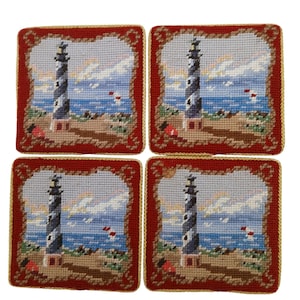 May include: A set of four square needlepoint coasters featuring a lighthouse scene. Each coaster displays a black and white striped lighthouse on a sandy beach with a blue ocean and sky, framed by a deep red border with gold piping trim.