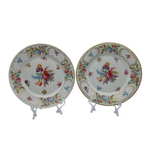 May include: Two white ceramic plates with floral designs. Each plate features a central bouquet of colorful flowers, including red, purple, and yellow blooms. The rims are adorned with gold trim and a decorative border. The plates are displayed on clear stands.