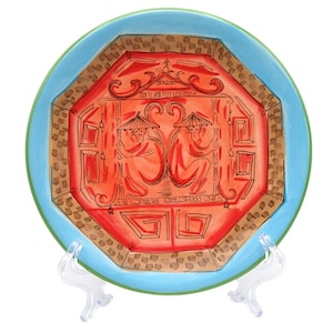 May include: A round ceramic plate featuring a central octagonal design in shades of red and brown. The illustration depicts two monkeys wearing conical hats, framed by geometric patterns and a bright light blue border with a thin green outer rim.