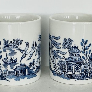 May include: Two white ceramic mugs with blue Willow pattern illustrations. The design features a pagoda, birds, and a boat on a lake. Each mug has a curved handle and a capacity of approximately 12 ounces.