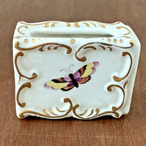 May include: A white porcelain box with a rectangular shape and a butterfly design. The butterfly has yellow, purple, and black wings. Gold scrollwork and accents adorn the edges of the box, which sits on a wooden surface.