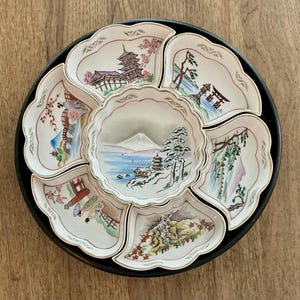 May include: A vintage Japanese serving set with a black tray and eight decorative porcelain dishes. Each dish features a different hand-painted landscape scene with pagodas, mountains, and trees. The dishes have scalloped edges and gold trim.