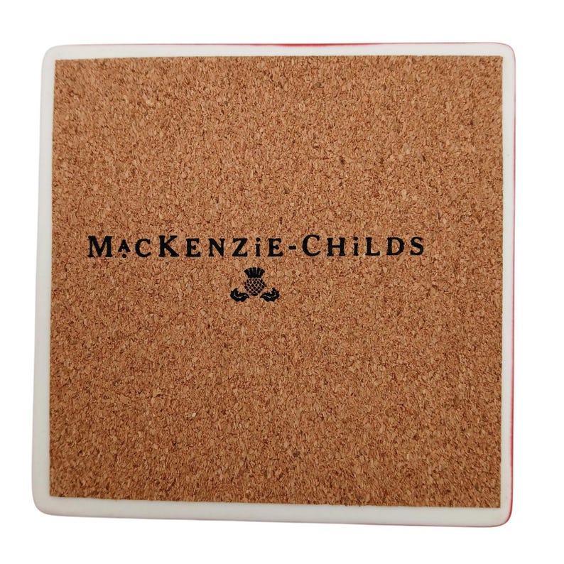 May include: A square coaster with a white border and a brown cork backing. The centre of the cork features the black text MACKENZIE-CHILDS printed above a small black thistle icon. The coaster has rounded corners and a smooth, flat surface.