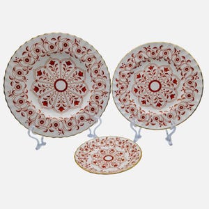 May include: Three white ceramic plates with a red floral design and gold trim. The plates are displayed on white stands. The largest plate has a diameter of approximately 10 inches, the medium plate is about 8 inches, and the smallest plate is about 6 inches.
