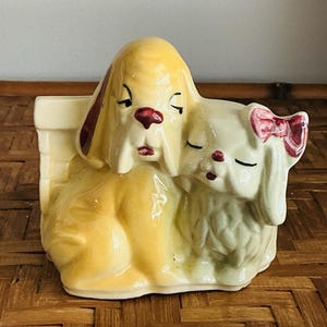 May include: A ceramic planter featuring two sleeping dog figurines. One dog is yellow with burgundy accents, and the other is off-white with a pink bow. The planter has a rectangular shape and is placed on a woven surface.