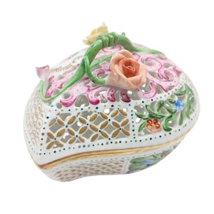 May include: A white porcelain trinket box with a pierced design and floral accents. The box features a lid adorned with pink and peach-colored roses, green vines, and delicate gold trim. The intricate lattice-like pattern adds to its decorative appeal.