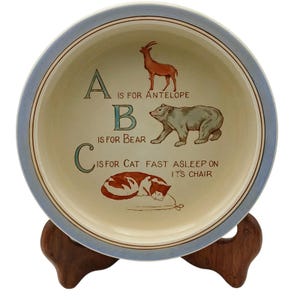 May include: A decorative ceramic plate with a light blue rim and a cream-colored center. The plate features the letters A, B, and C, along with illustrations of an antelope, a bear, and a sleeping cat. Text reads "A is for Antelope", "B is for Bear", and "C is for Cat fast asleep on it's chair".