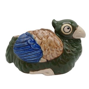 May include: A ceramic bird figurine featuring a glossy dark green body, a light beige beak, and large white eyes with black pupils. The wings are detailed with a textured pattern, painted in a combination of vibrant blue and light brown glaze.