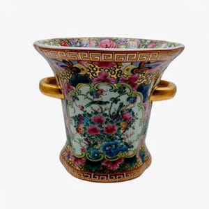 May include: A decorative porcelain vase with a flared top and two gold handles. The vase features a colorful floral design with pink, blue, and yellow flowers, and a gold geometric border. The vase is approximately 8 inches tall.