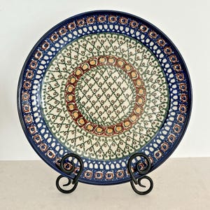 May include: A round ceramic plate with a blue rim and intricate floral patterns in red, green, and yellow. The plate is displayed on a black metal stand, showcasing its decorative design. The plate's diameter is approximately 10 inches.