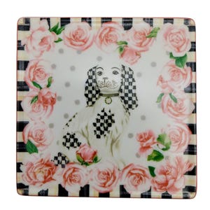 May include: A square ceramic plate featuring a central illustration of a white dog with black and white chequered ears and patches. The dog is surrounded by a border of pink roses on a white background with grey polka dots and black stripes.