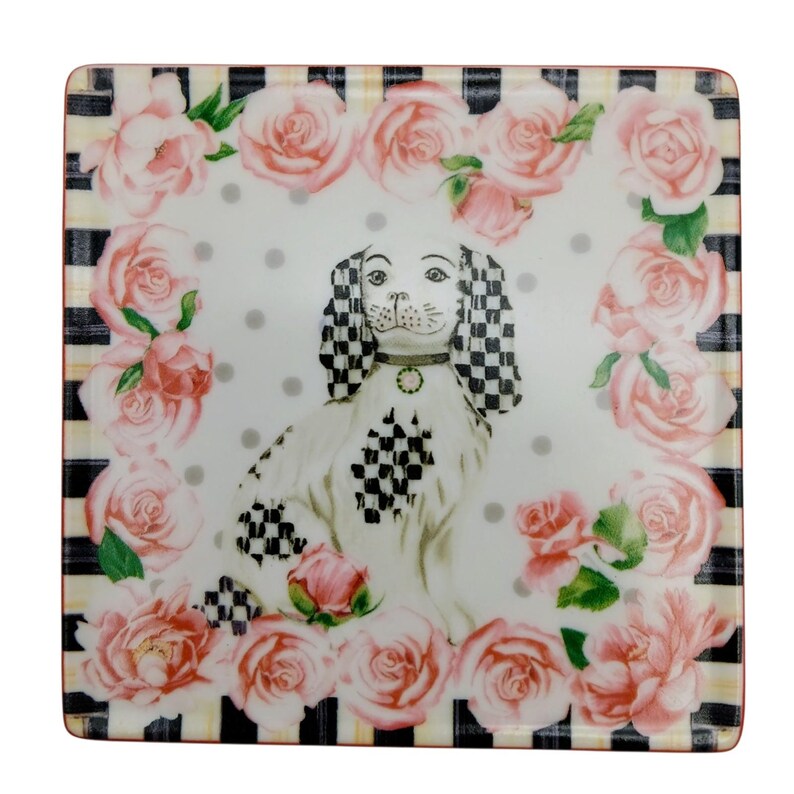May include: A square ceramic plate featuring a central illustration of a white dog with black and white chequered ears and patches. The dog is surrounded by a border of pink roses on a white background with grey polka dots and black stripes.
