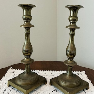 May include: Two antique brass candlesticks. Each candlestick has a square base, a slender, textured stem, and a flared top. The brass has a warm, aged patina, suggesting vintage or antique origins. The candlesticks are displayed on a white lace doily.