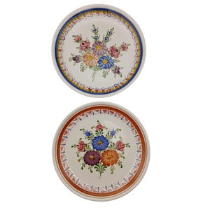 May include: Two white ceramic plates with hand-painted floral designs. Each plate features a central bouquet of colorful flowers, including pink, blue, purple, and orange blooms. The rims are decorated with intricate patterns in blue and red.