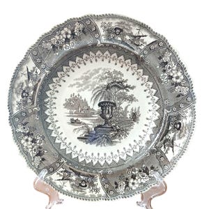 May include: A round ceramic plate featuring a black and white transferware pattern. The center depicts a landscape with a large urn, trees, and a boat on water. The border is decorated with floral motifs and small sailing ship vignettes.